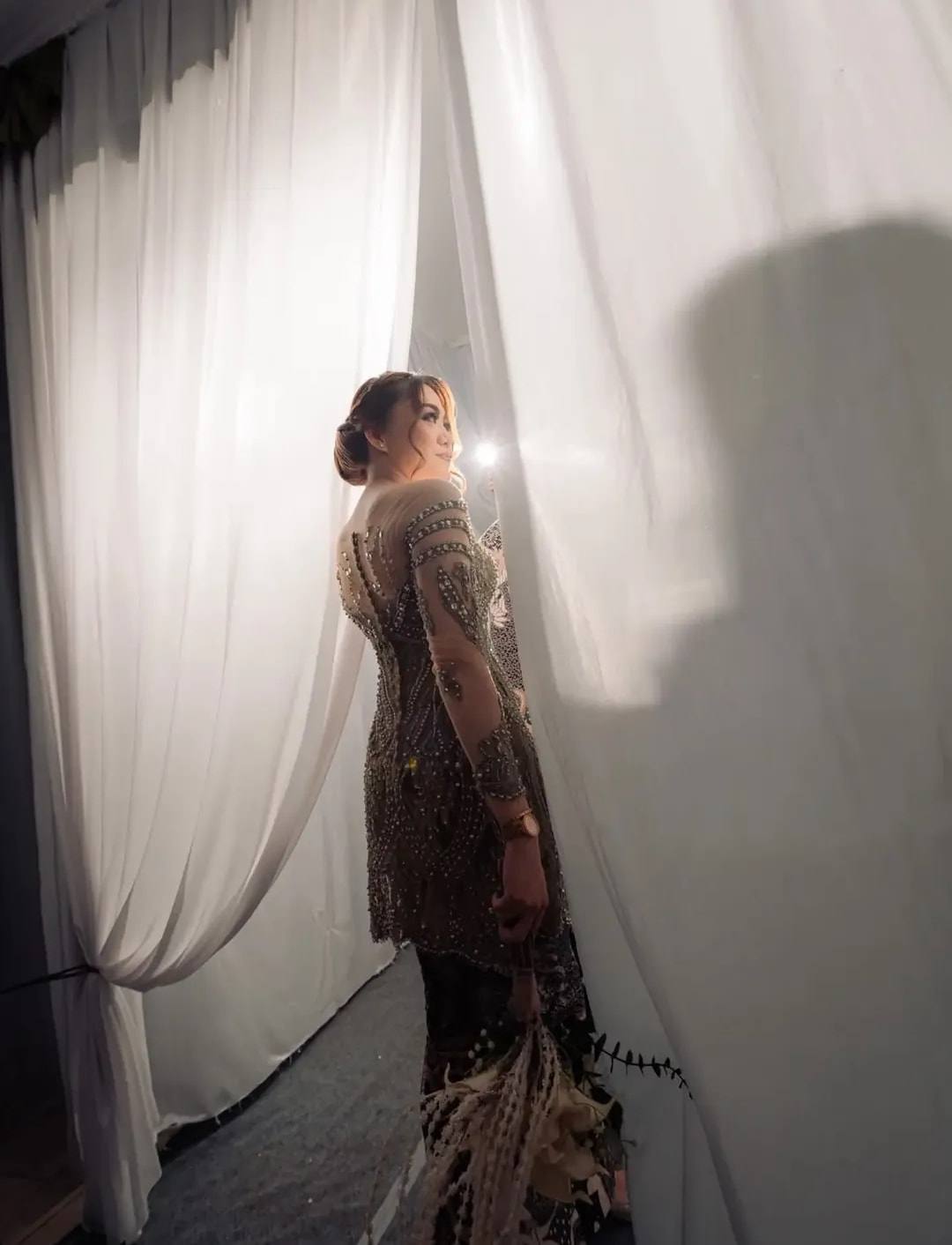 Detail punggung kebaya lamaran dengan rok berekor di depan tirai putih