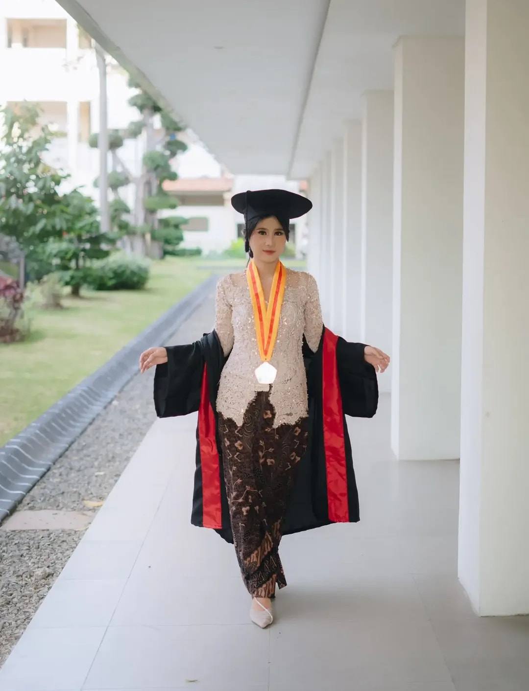 Wisudawati berkebaya krem memakai toga berjalan di lorong kampus