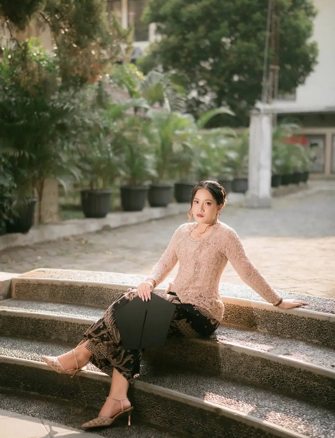 Kebaya renda merah muda dan kain batik gelap, wisudawati duduk di tangga taman