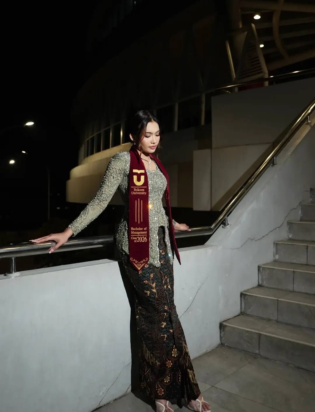 Kebaya sage dengan selempang wisuda di tangga gedung pada malam hari